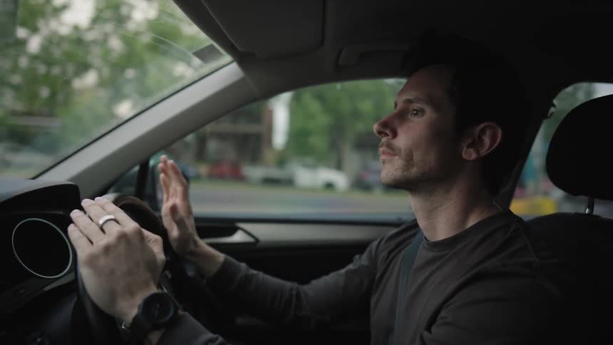A young man taps the steering wheel while driving in his car during the day