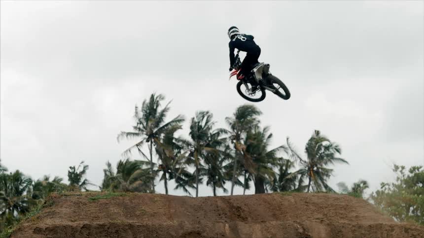 A person doing tricks on a motorbike on a dirt mound with palm trees in the back