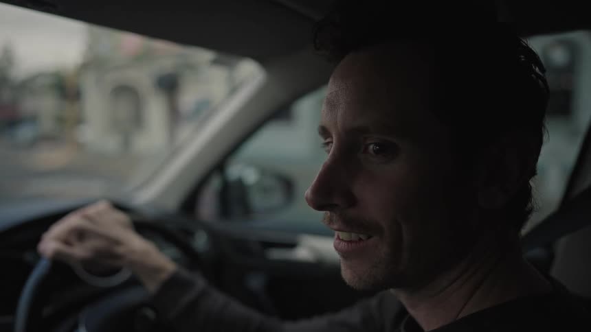 A man smiling slightly and talking while gripping the steering wheel of a car with the interior and street view slightly blurred in the background