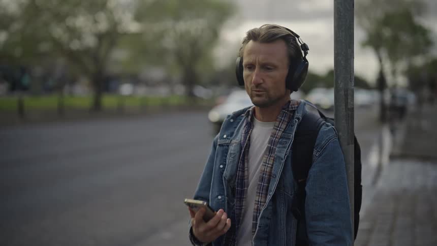 A man standing at a road sign by the highway listening to music with his headphones and holding the cellphone with one hand