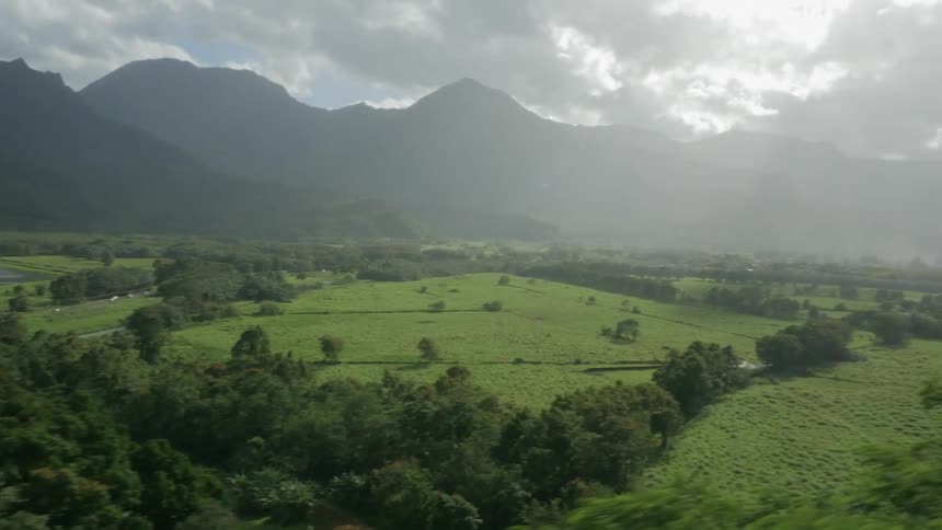 A rural area of trees and green grass at the bottom of mountains with many clouds in the sky