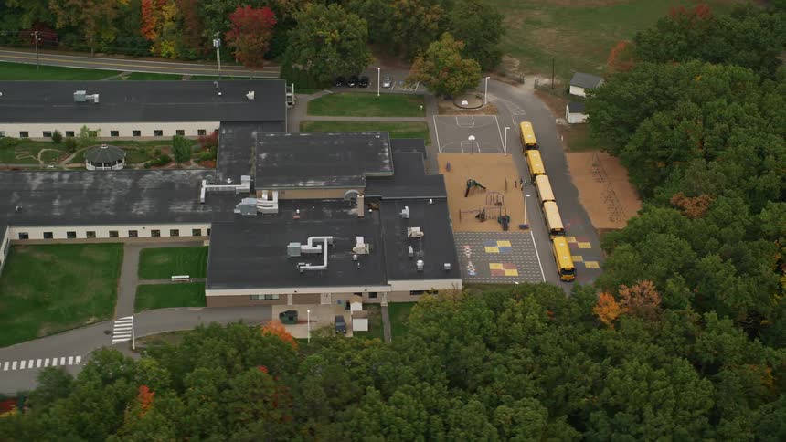 Buses park in a line in front of school surrounded by woods during fall