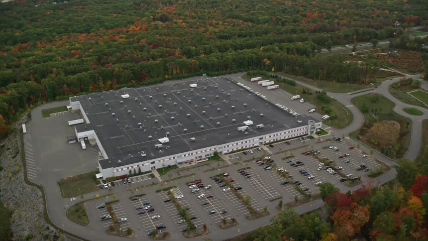 A large industrial building with a large car parking lot that is surrounded by forests