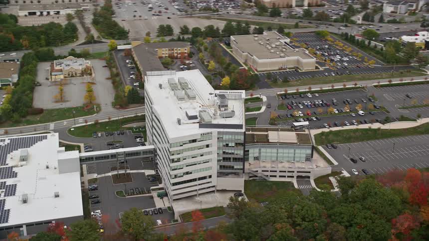 Aerial view of a modern office building with a large parking area surrounded by other commercial buildings parking lots and autumncolored trees