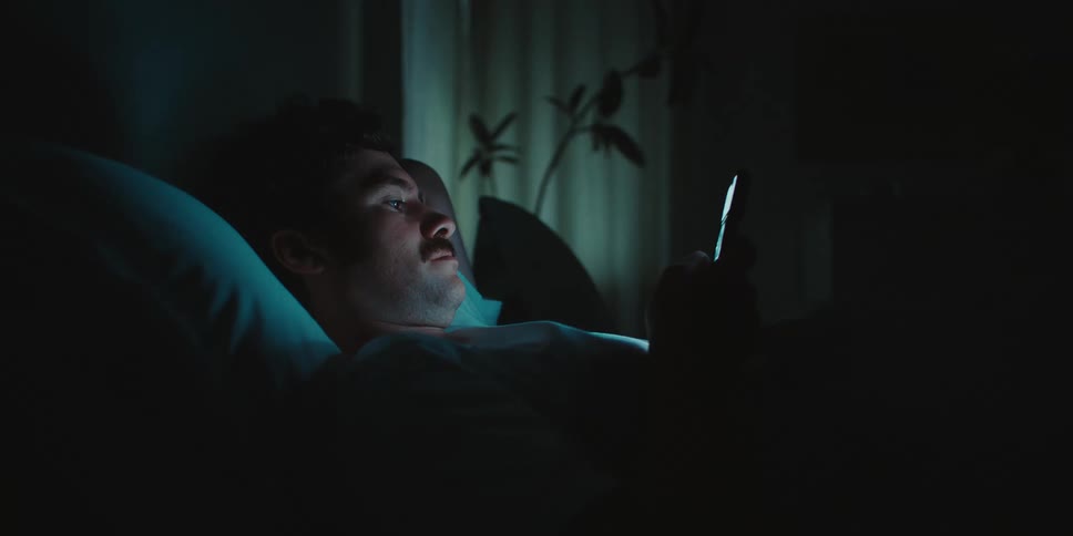 Man lying in bed at night looking at his smartphone with the screen illuminating his face