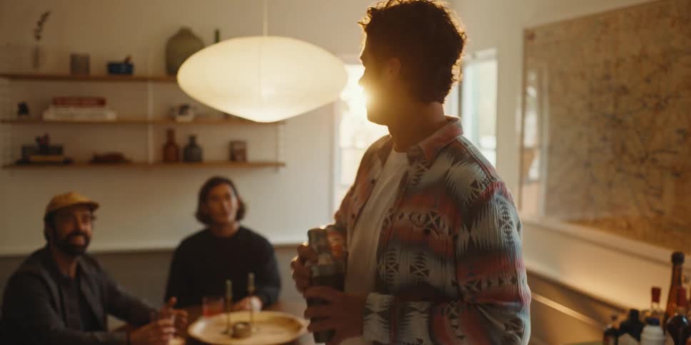 Man preparing an alcoholic drink with his hands while talking to his other two friends sitting at a table