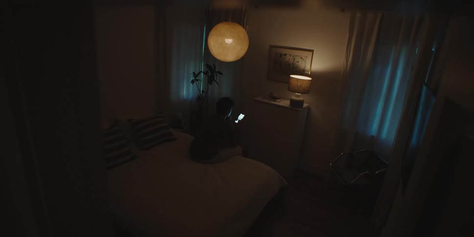 A man sits at the edge of his bed with his smartphone in a darkened room
