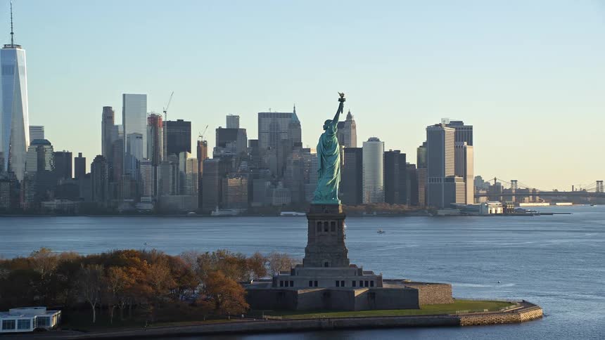 Aerial view of a statue of liberty on an island in the sea with a metropolis in the background