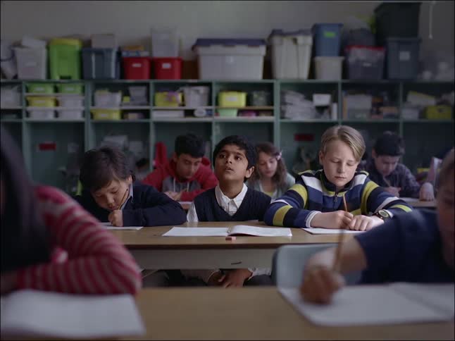 Children in a classroom writing and talking with one of them looking up thoughtfully