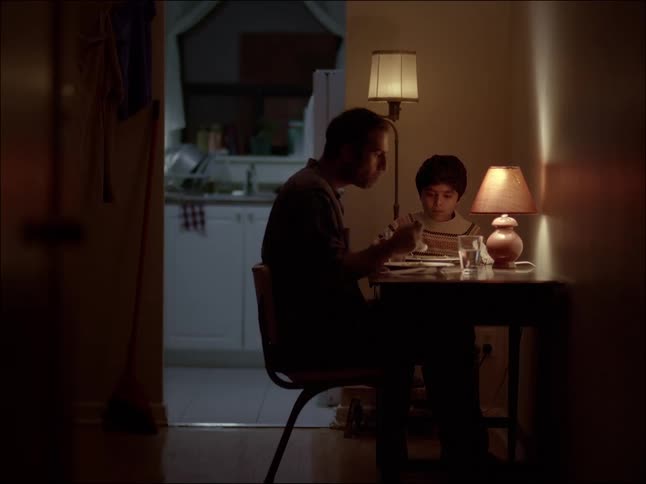 A man and a small boy are having dinner at a small table with light from a lamp