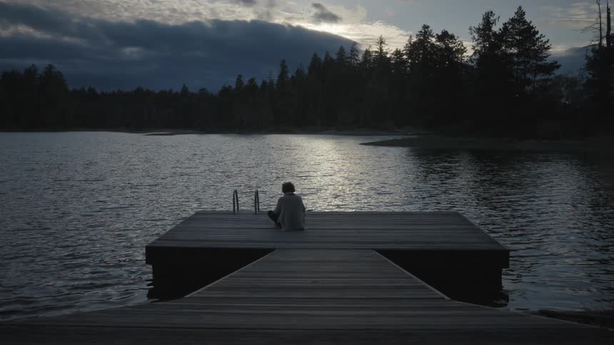 A man sitting on a dock facing a lake in slow motion