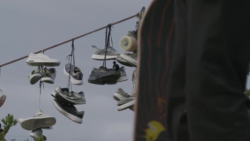 Several shoes hanging from a wire and a person with a skateboard passing in front