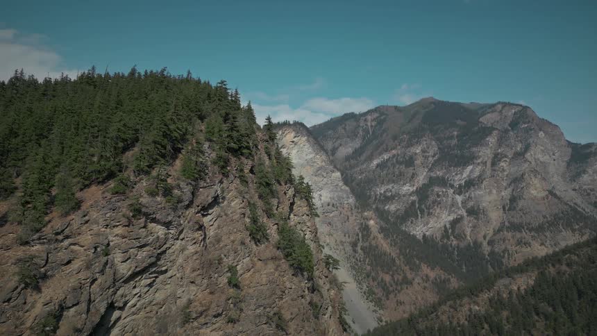 A region with some stone mountains and at the top of the closest one there is a pine forest