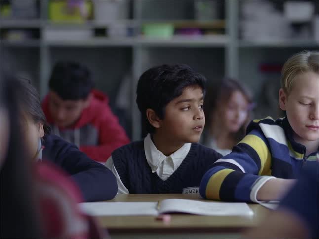 A boy in the classroom sitting in the middle of a desk with other students observing his classmates and looking thoughtful