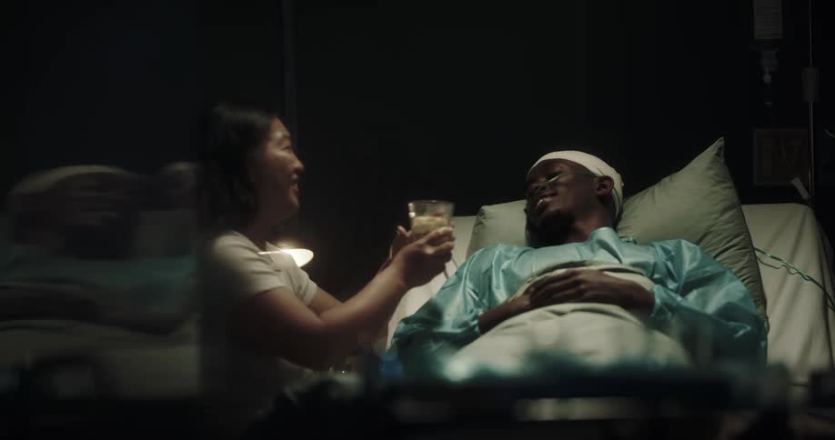 A woman talking to her partner who is lying in the hospital bed and offering him a glass of water
