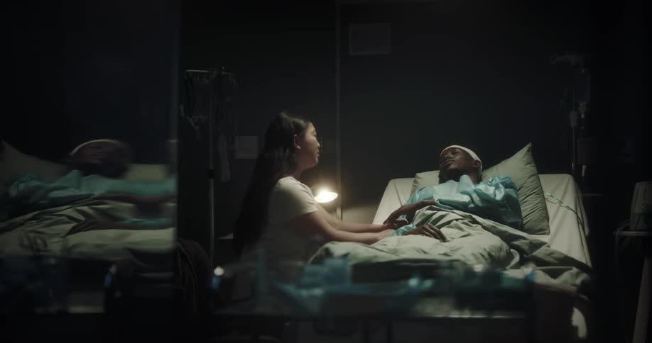 Woman caresses man lying in hospital bed in dim room