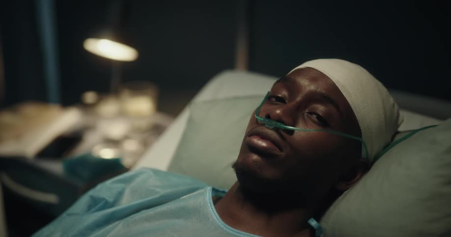 A young black man lies still in a hospital bed and looks around