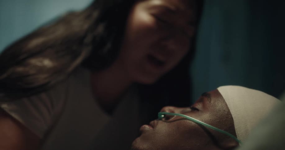 A woman crying and giving a kiss to her partner who is lying asleep on a hospital stretcher