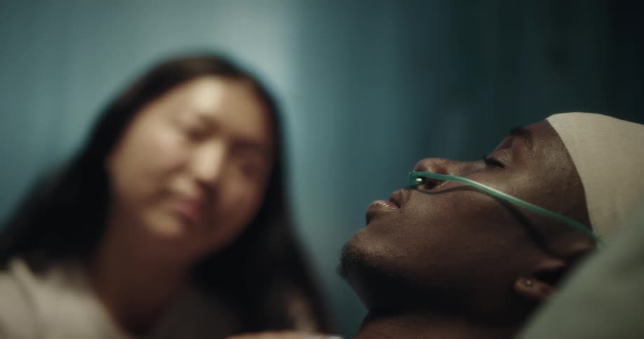 A woman affectionately tends to a man with a nasal cannula lying in a hospital bed