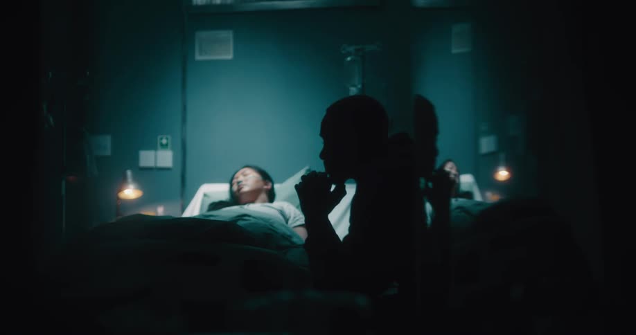 A black man walks up to a sleeping lady on the hospital bed and tucks her hand under the cover