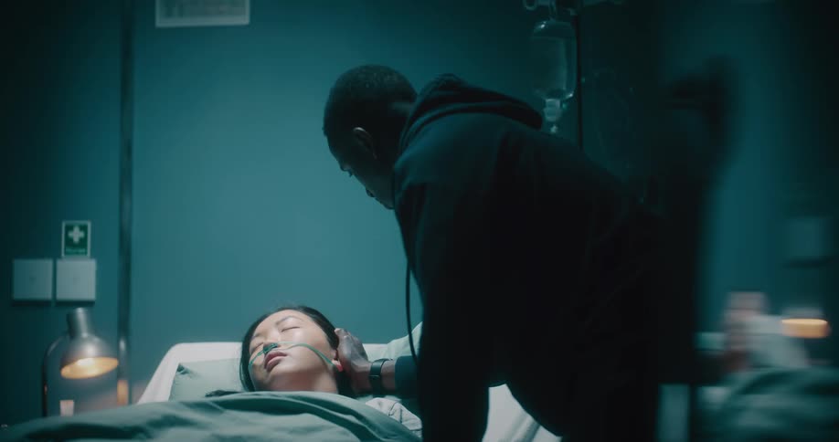 A black man kisses a sleeping asian lady on an hospital bed on the head