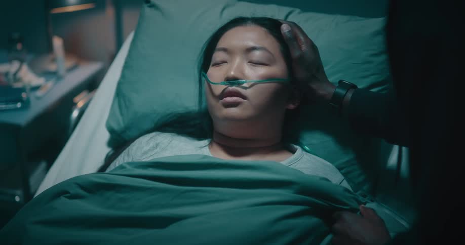 A woman with a breathing tube sleeps in a hospital bed as someone adjusts her sheets caresses her face and kisses her forehead