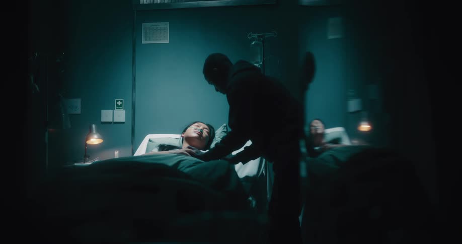 A young black man comforts a young asian woman while she lies in a hospital bed