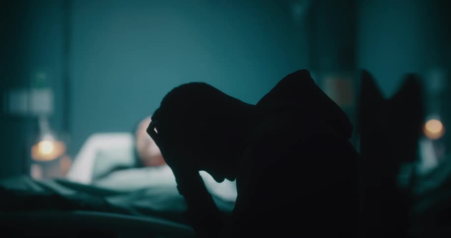 A sad man sitting in a chair next to his wife in the hospital bed