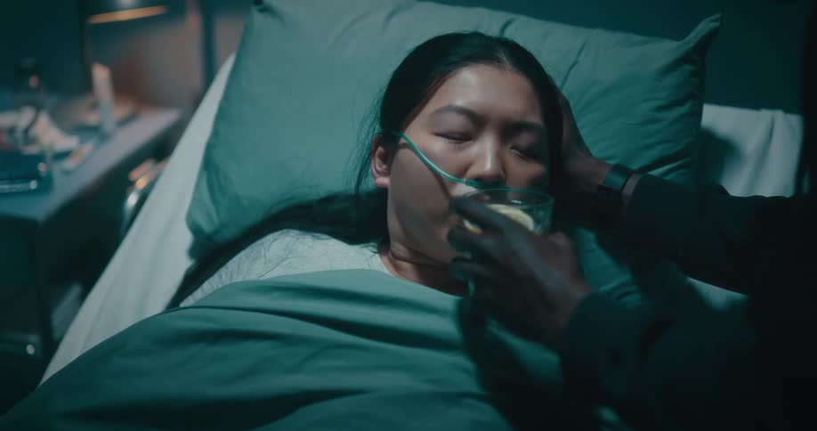 Man touches womans shoulder and gives her a glass of water lying in hospital bed