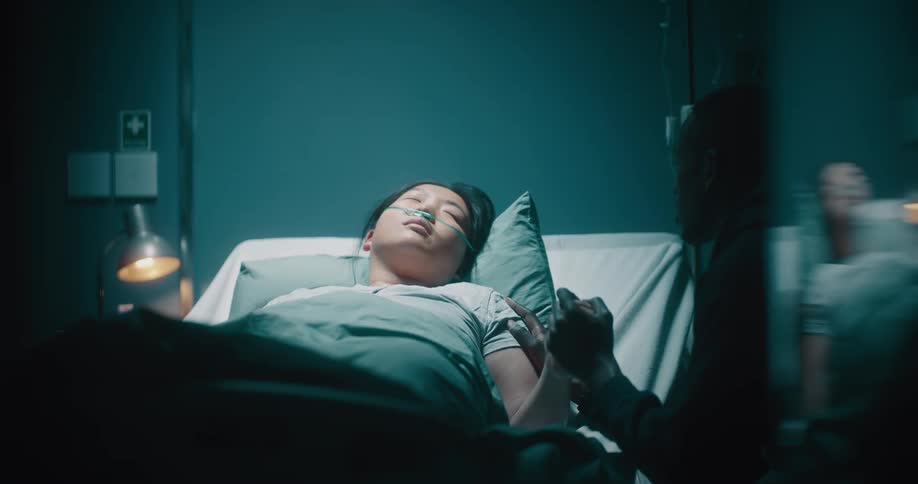A woman lies in a hospital bed and a man holds her hand