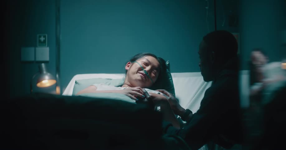 Asian lady on the hospital bed smiling with a man seated near the bed holding her hands