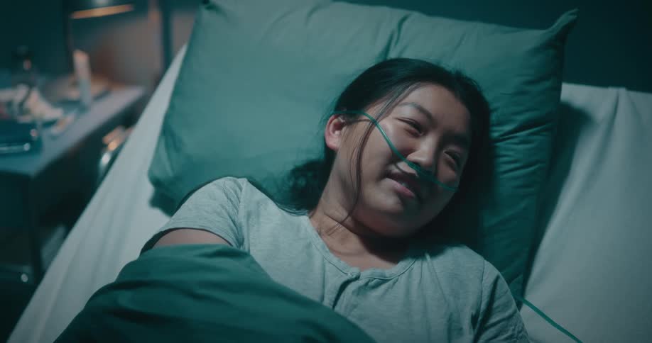 A woman in a hospital bed wearing a breathing tube smiles and laughs as she talks to someone