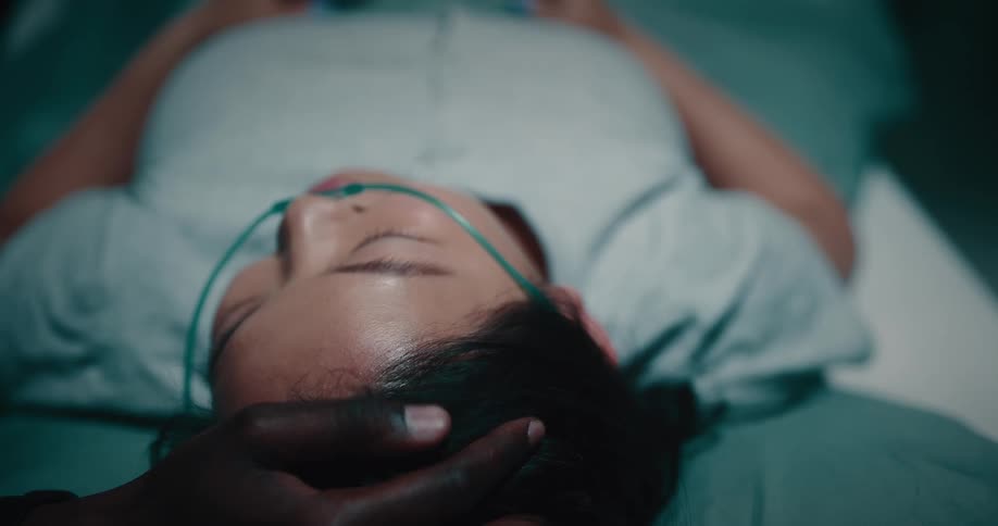 Lady lay on the hospital bed with tube in her nose while a black man rubs her head