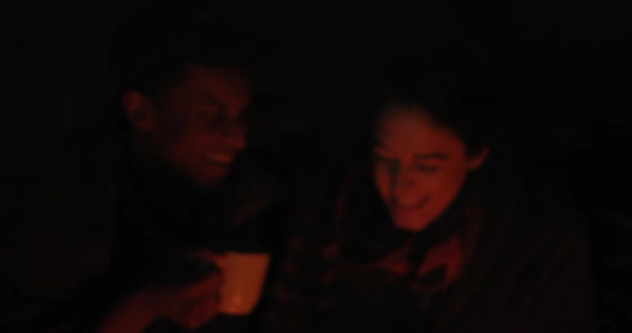 A man and a woman sitting by a campfire at night laughing and having a drink