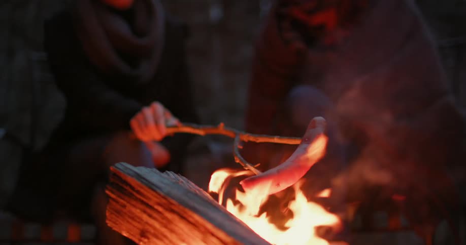 People sitting by a campfire at night and roasting a hot dog on a stick over the fire