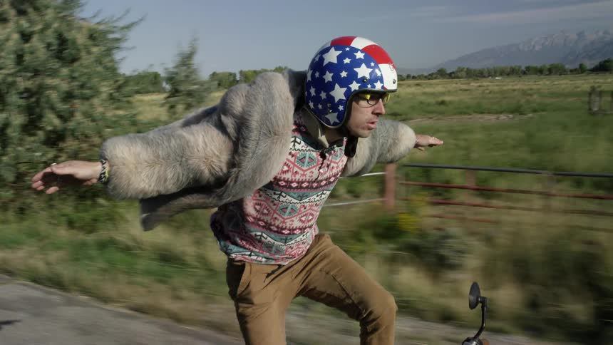 A man standing on a motor bike and riding it down a rural road with his arms outstretched