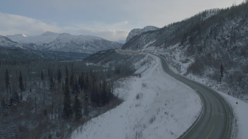 Ascending over an icy bridge deck to reveal a two lane highway that winds through snow capped mountains