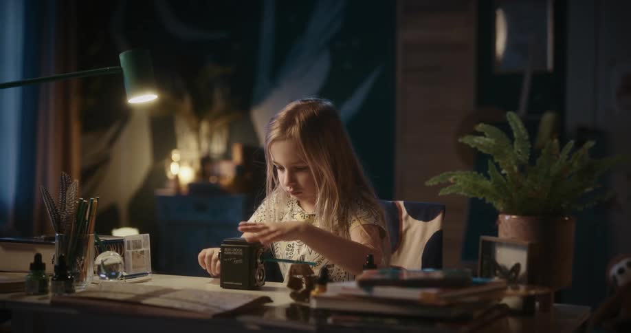 A young blonde girl sits at a desk and sharpens a pencil