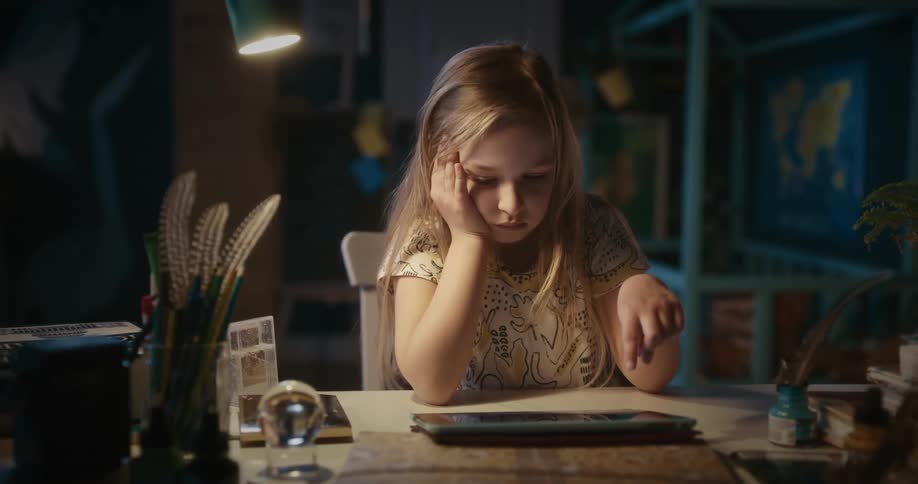 Girl playing with her tablet at a desk with her head resting on her hand in a warmly lit room