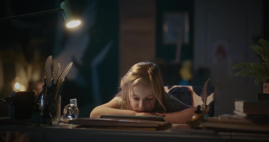 A little blonde girl has her arms and head on a table looking tired while watching something on a tablet