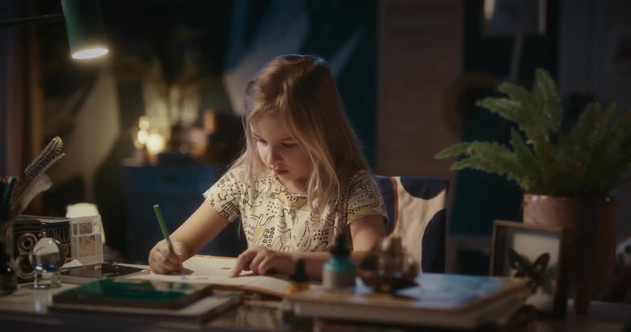 A little blonde girl is sitting and writing on paper with a pen