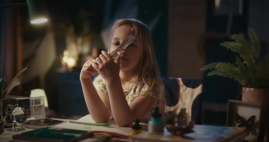 A girl sitting at a desk in a darkened room plays with a feather and smiles