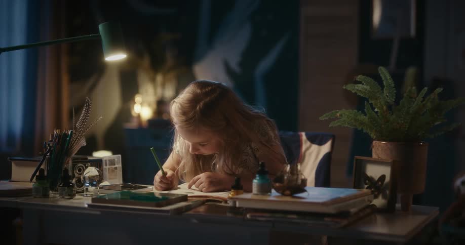 Girl sitting at table drawing looks up briefly with a smile