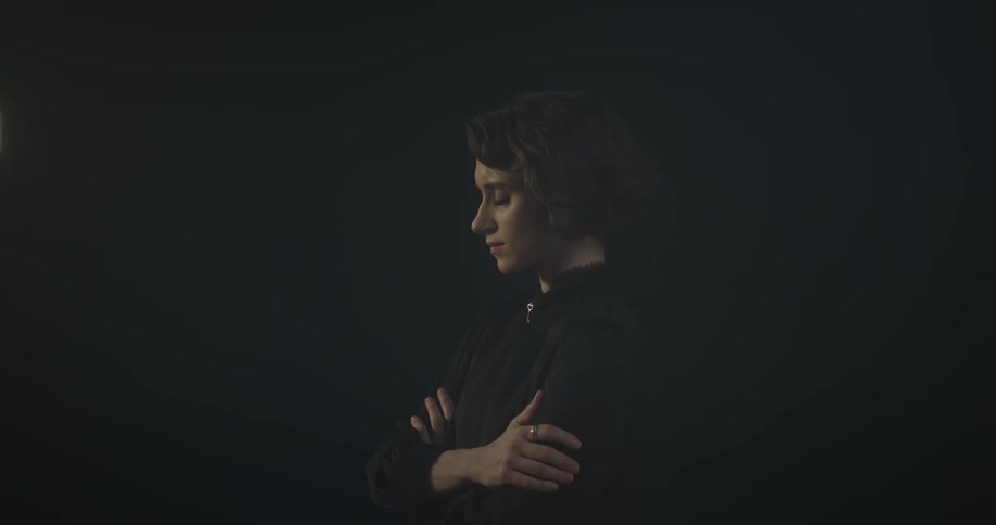 Woman with curly hair and a black shirt stands with her arms crossed in dark room