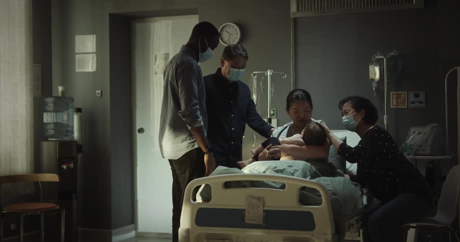 A black man gives a baby to the woman sitting on hospital bed with others greeting the woman