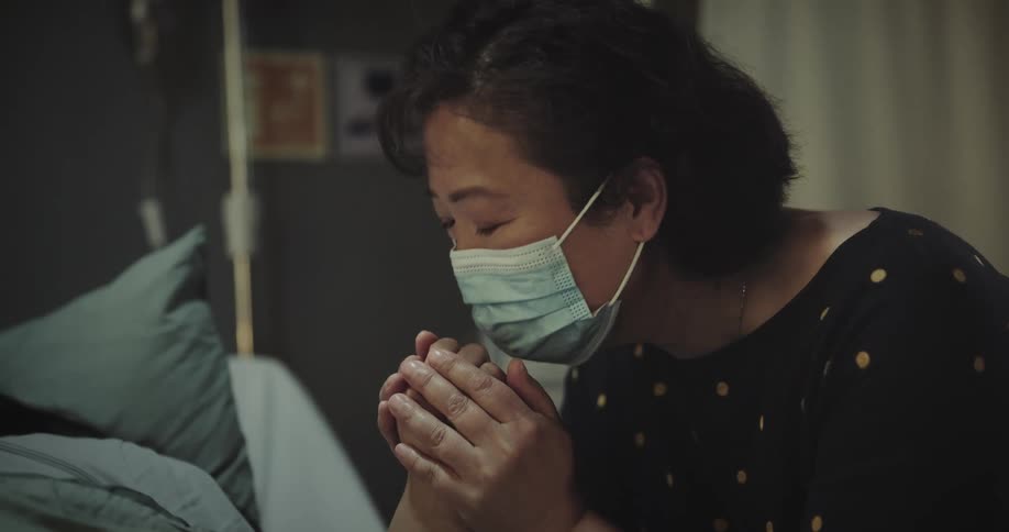 An elderly asian woman wearing a mask is holding a hospital patients hands and kissing them