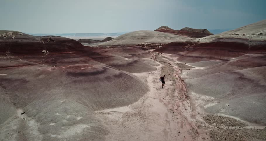 A man is hiking through rock layered mountains in the desert with mountains in the distance