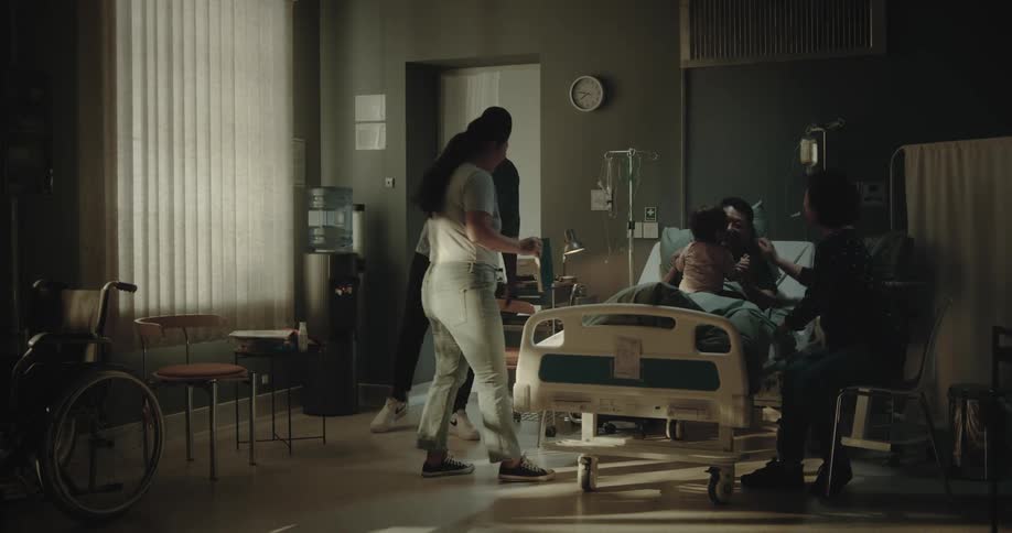 A man lying in a hospital bed reading a book puts the book away when 3 visitors bringing a small child arrive and sit in chairs next to his bed