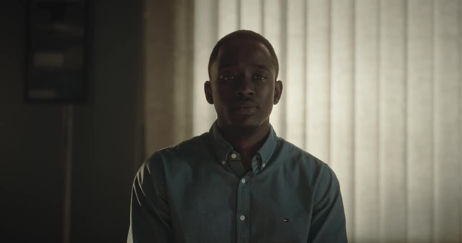 A man in a blue shirt looks directly at the camera with eye contact in a hospital room