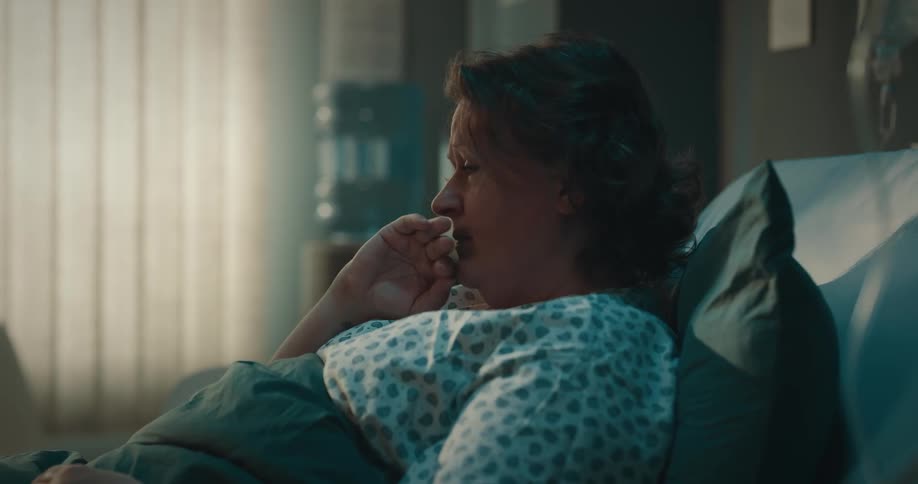 A woman lies in a hospital bed with a worried look and her hand on her face